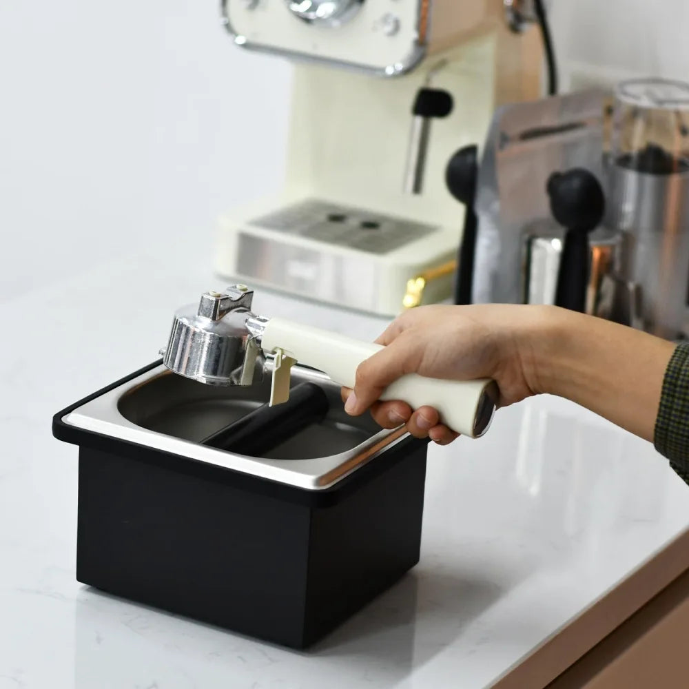 A barista holding an espresso portafilter and tapping it down onto the black silicone bar of the stainless steel dump bin to empty the coffee puck.