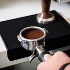 Perfectly leveled espresso coffee puck in stainless steel portafilter over black food-grade silicone mat.