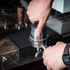 Barista pressing coffee grounds into portafilter on non-slip thick silicone edge mat protector.